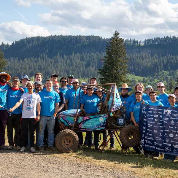 A group of CWRU Baja SAE members pose behind their competition vehicle, holding a sign featuring sponsor logos, in Oregon.